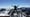 Silhouette d’une personne debout sur une plateforme en hauteur, bras ouverts face à un panorama de montagnes au-dessus des nuages.
